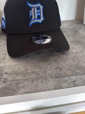 Svart New Era 9FORTY keps Detroit - Snygg svart keps från New Era, modell 9FORTY, med blått broderat D framtill för Detroit Tigers. Justerbar passform och böjd skärm. Klassisk basebollstil med broderade detaljer på sidorna. Perfekt accessoar för sportiga outfits. Aldrig använd!