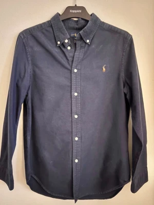 Mörkblå skjorta från Ralph Lauren - Klassisk mörkblå skjorta från Ralph Lauren med button-down krage och vita knappar. Skjortan har lång ärm och den ikoniska broderade loggan på bröstet. Tillverkad i bomull för en skön och stilren känsla. Storlek L i barn så ca s-m vuxen storlek. En ”half rare” skjorta.