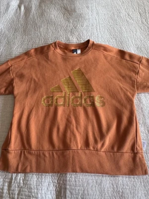 Orange Adidas sweatshirt med logga - Snygg orange sweatshirt från Adidas med stor guldig logga framtill. Tröjan har rund halsringning och är tillverkad i mjukt bomullsmaterial. Perfekt för en avslappnad och sportig stil.