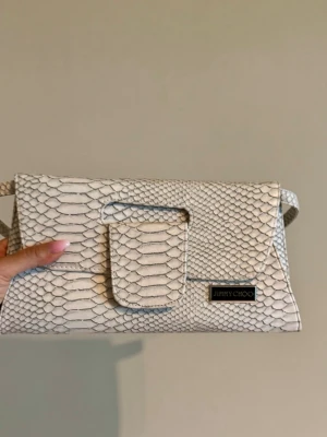 Vit kuvertväska från Jimmy Choo - Snygg vit kuvertväska i ett lyxigt ormskinnsmönstrat läder. Väskan har en stilren rektangulär form, ett litet lock framtill och en diskret logotyp i metall. Perfekt för dig som vill sticka ut med en trendig accessoar.