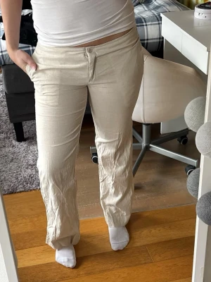 Beiga linnebyxor med raka ben - Snygga beiga linnebyxor med raka ben och klassisk knappgylf. Byxorna har normal midja och sidfickor, perfekta för en avslappnad och trendig look. Tillverkade i ett lätt och luftigt linnematerial som passar bra till varmare dagar.