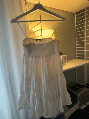 Vit sommarklänning med smock - Luftig vit klänning med smala axelband och smock upptill. Kjolen är volangig och har en avslappnad, flowy passform. Perfekt för varma dagar och enkel att styla med accessoarer. Tillverkad i ett tunt, skrynkligt tyg som ger en bohemisk känsla. Axelbandet har gått av, så jag satt dit en säkerhetsnål