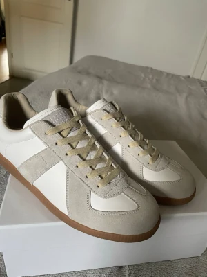 Vita och beige sneakers med mockadetaljer - Snygga sneakers i vitt och beige med ovandel i läder och mocka, samt klassisk gummisula. Skorna har beige snörning och en stilren, retroinspirerad design som passar till det mesta. Perfekta för dig som gillar minimalistisk och clean stil.