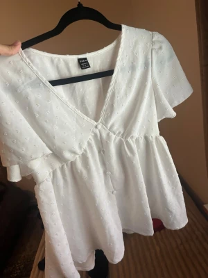 Vit blus med v-ringning från Shein - Söt vit blus från Shein med v-ringning och korta ärmar. Blusen har små broderade detaljer och knappar framtill. Den har en luftig passform och lätt volang nedtill, perfekt för en fräsch och somrig look.