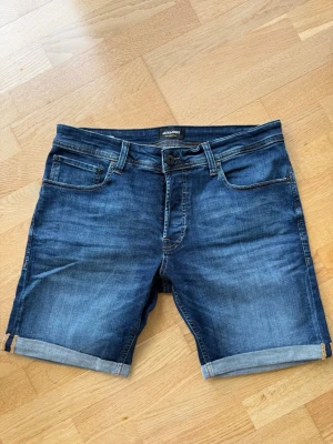 Mörkblå jeansshorts från Jack & Jones - Snygga mörkblå jeansshorts från Jack & Jones med klassisk femficksdesign och uppvikta benslut. Tillverkade i denim med en lätt tvättad look och detaljerade sömmar. Perfekta för en avslappnad stil under varma dagar.