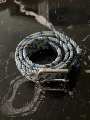 Flätat blått bälte från ONEWear - Snyggt flätat bälte från ONEWear i olika blå nyanser, beige och vitt. Bältet har detaljer i mörkt skinn och ett klassiskt metallspänne. Perfekt accessoar för att lyfta din outfit med en cool touch.