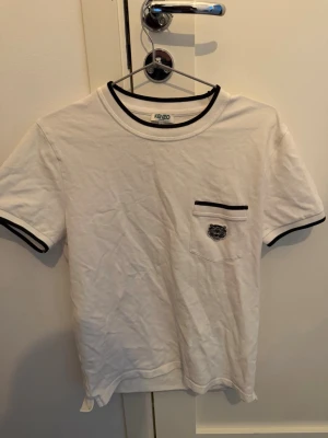 Vit Kenzo t-shirt med svart kant - Vit t-shirt från Kenzo med svarta detaljer vid krage, ärmslut och bröstficka. Klassisk passform och liten Kenzo-logga på fickan. Perfekt för en stilren och trendig look.