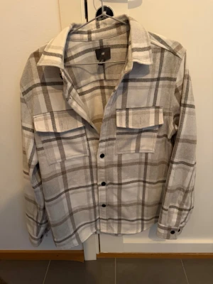 Beige och vit rutig overshirt H&M - Overshirt från H&M i beige och vitt rutigt mönster. Skjortjackan har två bröstfickor med lock, knäppning framtill och klassisk krage. Tillverkad i mjukt tyg som ger en avslappnad look. Perfekt lager på lager-plagg för en trendig stil.