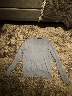 Ljusblå  tröja från Ralph Lauren - Snygg ljusblå  tröja från Ralph Lauren med rund halsringning och diskret broderad logga på bröstet. Tillverkad i mjukt material och har långa ärmar samt ribbade muddar vid ärmslut och nederkant. Perfekt för en clean och stilren look.