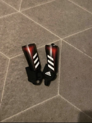 Adidas benskydd svart/röd - Säljer ett par Adidas benskydd i svart och rött med vita detaljer och klassiska tre ränder. Skydden har elastiska band och tålig yta, perfekta för fotbollsträning eller match. Adidas-logga framtill och bekväm passform.
