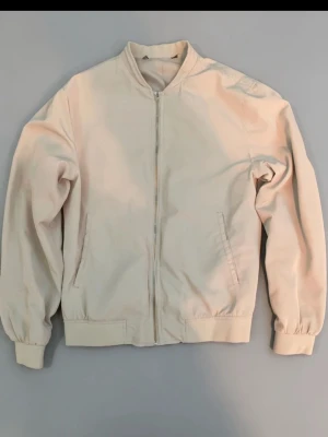 Beige bomberjacka med dragkedja - Stilren beige bomberjacka med klassisk passform, ribbade muddar och dragkedja framtill. Jackan har två snedställda fickor och är tillverkad i ett mjukt tyg som ger en clean look. Per dig som gillar minimalistisk stil.
