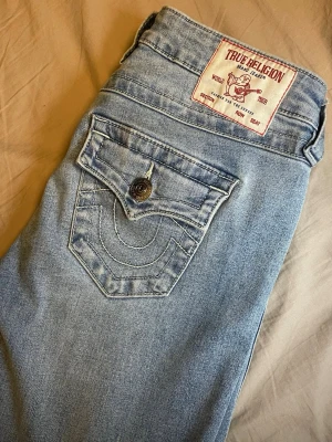 Ljusblå bootcut jeans från True Religion - Säljer ett par ljusblå bootcut jeans från True Religion med ikoniska bakfickor och snygga kontrastsömmar. Måste tyvärr sälja dem pga stor storlek. Har bara använt några gånger men har behövt ha bälte. Dem är i nytt och jättebra skick. Storlek är 25 vilket jag räknat ut är runt S-XS. Jag brukar ha storlek XS, dem är långa i benen och måste stoppa in dem i skorna för att inte trampa på dem. är runt 167cm lång. 