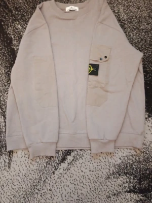 Beige sweatshirt från Stone Island - Snygg beige sweatshirt från Stone Island med klassisk logotyp på ärmen och en bröstficka med knappdetaljer. Tröjan har rund halsringning och är tillverkad i mjukt material som passar perfekt till en avslappnad stil.