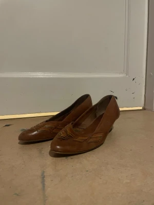 Bruna klassiska ballerinaskor i läder - Snygga bruna ballerinaskor i äkta läder med flätade detaljer framtill. Skorna har en spetsig tå och låg klack, vilket ger en tidlös och elegant look. Perfekta för dig som gillar vintagevibbar och klassisk stil.