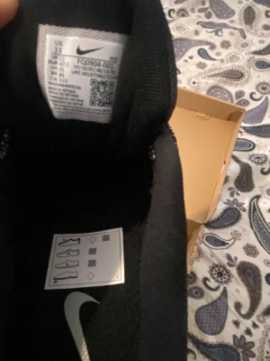 Nike Adapt Auto Max svarta sneakers - Nike Adapt Auto Max sneakers i svart med grå detaljer och vit sula. Skorna har en futuristisk design med mesh- och syntetmaterial, samt en synlig Nike-logga på sidan. De har en chunky sula och smart snörning för en modern look.