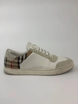 Burberry vita låga sneakers - Snygga vita sneakers från Burberry med klassiskt rutigt mönster på hälen och beige mockadetaljer. Skorna har vita skosnören, perforerade detaljer på sidan och en låg silhuett. Tillverkade i en mix av läder och mocka för en exklusiv känsla.