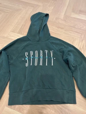 Grön hoodie SPORTY från Gina Tricot - Mörkgrön hoodie från Gina Tricot med trycket 'SPORTY' i vitt framtill. Hoodien har huva och ribbade muddar. Perfekt för en avslappnad och sportig stil. Mjuk insida för extra komfort.