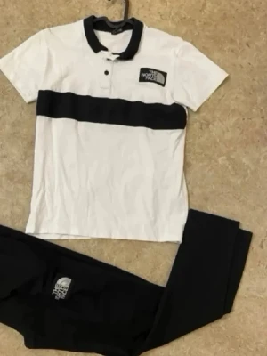 The North Face svartvitt set - Snyggt set från The North Face med vit pikétröja med svarta detaljer och matchande svarta byxor. Tröjan har svart krage, knappslå och logga på bröstet. Byxorna är helsvarta med logga. Perfekt för en sportig och avslappnad look.