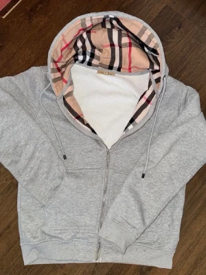 Grå hoodie med mönstrad huva - Snygg grå zip-hoodie från Burberry med klassiskt rutigt foder i huvan. Tröjan har dragkedja framtill, dragsko i huvan och ribbade muddar vid ärmslut och nederkant. Perfekt för en chill och stilren look.
