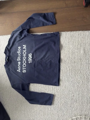 Mörkblå långärmad topp Acne Studios - Mörkblå långärmad topp från Acne Studios med vit text framtill: 'Acne Studios STOCKHOLM 1996'. Klassisk rak passform och rund halsringning. Perfekt för en stilren och avslappnad look.