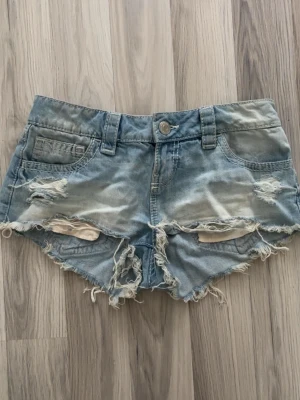 Ljusblå jeansshorts från Gina14+ - Säljer ett par ljusblå jeansshorts med råa kanter och slitna detaljer framtill. Shortsen har fem fickor, bälteshällor och knappgylf. Perfekta för sommaren och ger en avslappnad vibe. Storlek xs men passar s