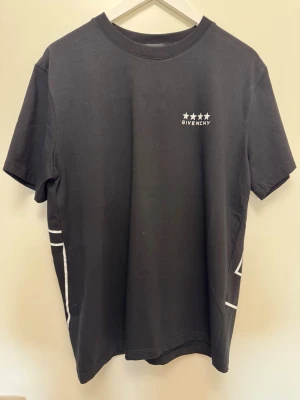 Svart t-shirt med grafiskt tryck - Svart t-shirt med vit logga och stjärnor på bröstet samt stort grafiskt mönster på ryggen. Klassisk rund hals och korta ärmar. Tillverkad i mjuk bomull för en skön känsla och snygg passform.