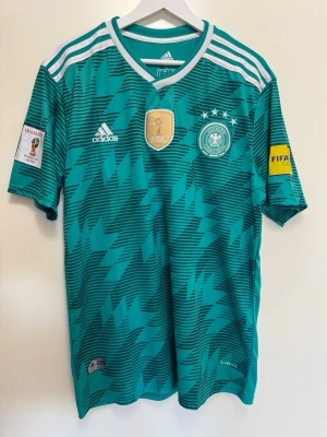 Tysklands fotbollströja Adidas grön - Grön Tyskland fotbollströja från Adidas med vita detaljer och mönster i svart. Tröjan har officiella FIFA och VM-märken, samt broderad landslagslogga och fyra stjärnor. Korta ärmar och rund hals. Tillverkad i lätt och ventilerande material.