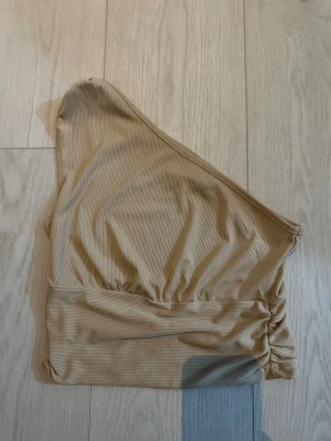 Beige ribbad bikiniöverdel - Snygg beige bikiniöverdel med ribbad struktur och asymmetrisk, one shoulder-design. Perfekt för strandhäng eller poolparty. Stilren och modern look som passar till det mesta.