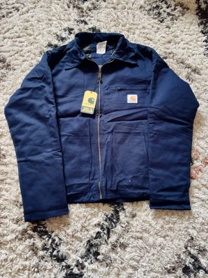 Mörkblå worker jacka från Carhartt - Snygg mörkblå overshirt från Carhartt med dragkedja framtill och klassisk krage. Bröstficka med Carhartt-logga och en extra ficka med dragkedja. Tillverkad i slitstarkt material, perfekt för lager-på-lager och streetwear.
