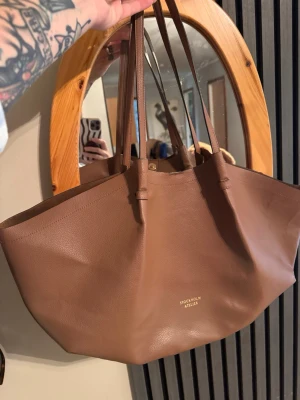 Brun shoppingväska från Stockholm Atelier - Snygg och rymlig shoppingväska i brunt läder från H&M  Stockholm Atelier. Väskan har två långa axelband och stängs med magnetknapp. Minimalistisk design med diskret logga framtill. Perfekt för dig som gillar stilrena accessoarer.