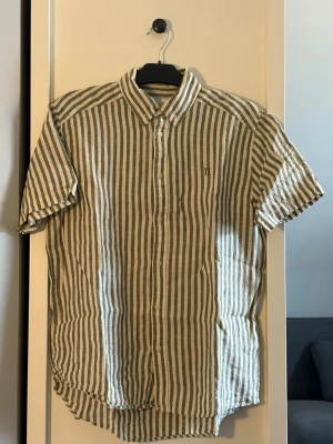 Randig kortärmad linneskjorta från les deux - Snygg kortärmad skjorta från Les Deux i beige och vit med vertikala ränder. Skjortan är tillverkad i linne, har klassisk krage och knäppning framtill. Perfekt för varma dagar och en avslappnad stil. Nypris 1099kr.