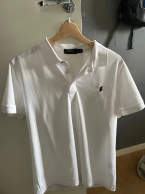 Vit pikétröja från Polo Ralph Lauren - Klassisk vit pikétröja från Polo Ralph Lauren med korta ärmar och krage. Tröjan har knappar framtill och den ikoniska broderade loggan på bröstet. Tillverkad i bomull för en skön och luftig känsla.