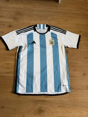 Argentina fotbollströja VM 2022 - Argentina fotbollströja från VM 2022 med blå och vita vertikala ränder, svarta detaljer och AFA-emblem på bröstet. Tröjan har korta ärmar, rund hals och är tillverkad i lätt funktionsmaterial. Perfekt för fotboll eller träning. Pris kan diskuteras vid snabb affär.