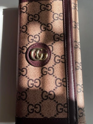 Plånbok brun gg - Snygg plånbok från gg. Utsidan är beige med mörkbrunt GG-tryck och insidan är i brunt läder med flera kortfack. Plånboken har guldfärgad logodetalj och är tillverkad i canvas och läder.