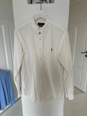 Vit skjorta från Polo Ralph Lauren - Klassisk vit slim fit skjorta från Polo Ralph Lauren med button-down krage och broderad marinblå logga på bröstet. Skjortan har långa ärmar, knappar framtill och är tillverkad i mjuk bomull. Perfekt till en clean och stilren look. 