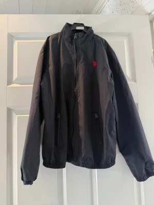 Svart vindjacka från U.S polo Assn. - Snygg svart vindjacka från U.S Polo Assn. med dragkedja framtill, två fickor med dragkedja och elastiska muddar. Jackan har en röd broderad logga på bröstet och står emot vind bra. Perfekt för blåsiga dagar.