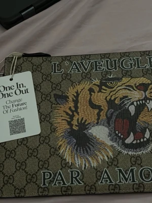 Hand  kuvertväska med tigertryck - Kuvertväska  i beige canvas med klassiskt GG-mönster, broderad tiger och texten 'L'Aveugle Par Amour' på framsidan. Svart avtagbart handledsband i läder och dragkedja upptill. Perfekt för att sticka ut med en lyxig touch.