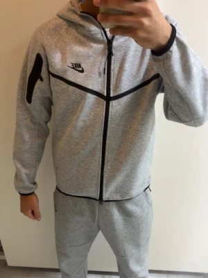 Grå Nike loungewear set - Snyggt grått loungewear set från Nike med dragkedja framtill och svarta detaljer.ENDAST ANVÄNT EN GÅNG, Säljer för att de inte är min stil.  Setet består av en hoodie med ficka på ärmen och matchande byxor. Tillverkad i mjukt och bekvämt material, perfekt för chill dagar hemma.