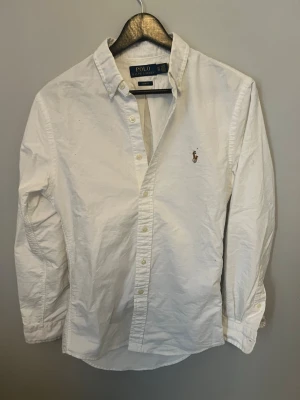 Vit skjorta från Polo Ralph Lauren - Klassisk vit skjorta från Polo Ralph Lauren i slim fit-modell. Skjortan har knappar framtill, lång ärm och den ikoniska broderade loggan på bröstet. Tillverkad i bomull och passar perfekt till en stilren look.