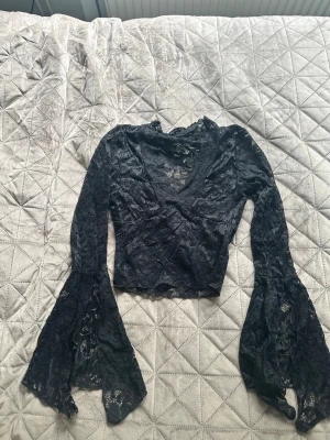 Svart spetsblus med vida ärmar - Snygg svart blus i spets med v-ringning och vida, utsvängda ärmar. Croppad modell som ger en trendig look. Perfekt för dig som vill sticka ut med coola detaljer och transparent material. Tajtare runt armarna