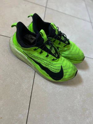 Nike ZoomX Vaporfly NEXT% löparskor - Säljer ett par Nike ZoomX Vaporfly NEXT% löparskor i neonlime med svarta detaljer och stor svart Swoosh på sidan. Ovandelen är i mesh för bra ventilation och sulan är tjock och responsiv för maxad löpkänsla. Skorna har snörning och en sportig, modern design.