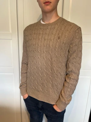 Ljusbrun kabelstickad tröja från Polo Ralph Lauren - Säljer en ljusbrun kabelstickad tröja från Polo Ralph Lauren med rund hals och diskret logga broderad på bröstet. Tröjan har långa ärmar och ribbade muddar vid ärmslut och nederkant. Perfekt för dig som gillar klassisk och stilren design. Det står att den är i XL men jag skulle säga att den sitter som L