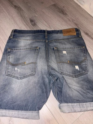 Blå jeansshorts med slitningar - Snygga blå jeansshorts från jack & Jones vintage med slitna detaljer och uppvikta benslut. Klassisk femficksmodell med normal passform och läderpatch bak i midjan. Perfekta för en avslappnad stil.