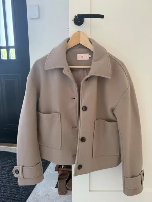 Beige jacka från Nelly -  Beige jacka från Nelly. Jackan har stora framfickor, klassisk krage, knäppning med knappar och snygga detaljer vid ärmsluten. 