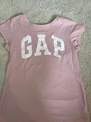 Rosa GAP t-shirt med glittrig text - Säljer en ljusrosa t-shirt från GAP med stort vitt glittrig logotryck på bröstet. T-shirten har korta ärmar och rund halsringning. Perfekt för en avslappnad och trendig look.