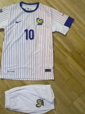 Frankrike fotbollströja Mbappé #10 - Säljer en vit fotbollströja med blå och röda ränder från Nike, inspirerad av Frankrikes landslag. Tröjan har blå detaljer, nummer 10 och Mbappé på ryggen samt tupp-logga på bröstet. Matchande vita shorts med samma logga ingår.