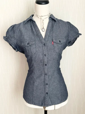 Blå kortärmad prickig  denimskjorta från Levi's - Snygg blå skjorta från Levi's med diskret mönster, korta puffärmar och två bröstfickor med knappar. Skjortan har figurnära passform, klassisk krage och knäppning framtill. Perfekt för en trendig och avslappnad look.