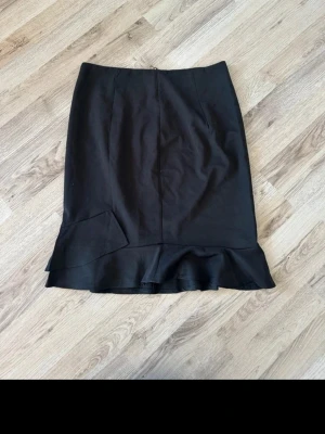 Svart kjol med volangkant - Snygg svart kjol med volangdetalj längst ner. Kjolen har en rak passform och en diskret söm framtill. Perfekt för dig som gillar stilrena plagg med en twist. Ingen synlig logga eller märke.