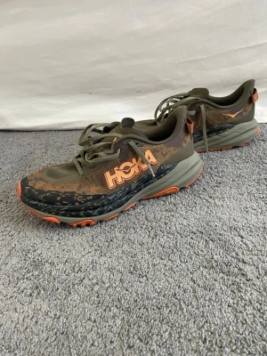 Hoka gröna löparskor med orange detaljer - Säljer ett par Hoka speedgoat 6 löparskor i grönt med orange detaljer och mönstrad sula. Skorna har mesh-ovandel för bra ventilation, klassisk snörning och en rejäl, dämpad sula som ger skön komfort vid löpning eller träning.
