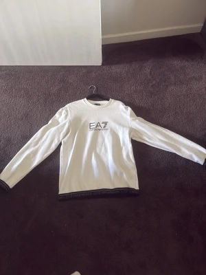 Vit EA7 Emporio Armani sweatshirt - Snygg vit sweatshirt från EA7 Emporio Armani med svart logga på bröstet och svarta detaljer med märkets namn längs nederkanten. Tröjan har rund hals och långa ärmar, perfekt för en stilren och sportig look.